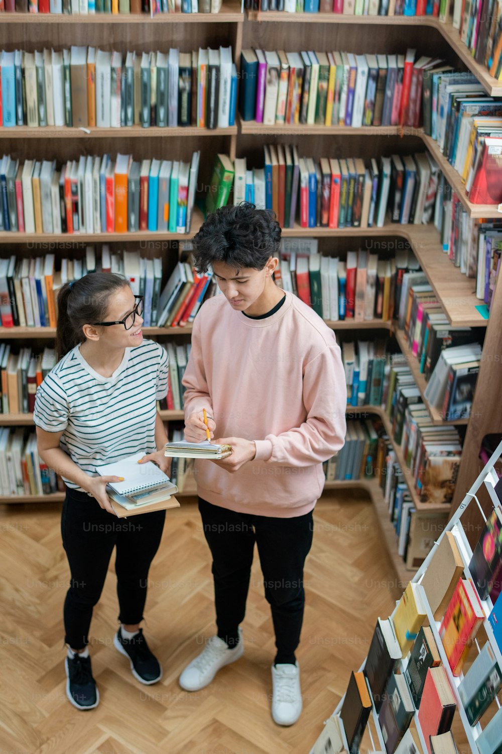 Two casual teenagers standing in college library among shelves with books and discussing home task after classes