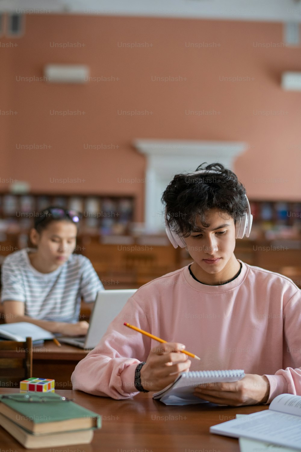 Ernsthafter Student mit Kopfhörern, der Musik hört, während er Notizen im Notizblock am Schreibtisch mit Mädchen im Hintergrund liest