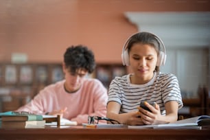 Pretty college student with headphones scrolling in smartphone while sitting by desk in reading hall of college library
