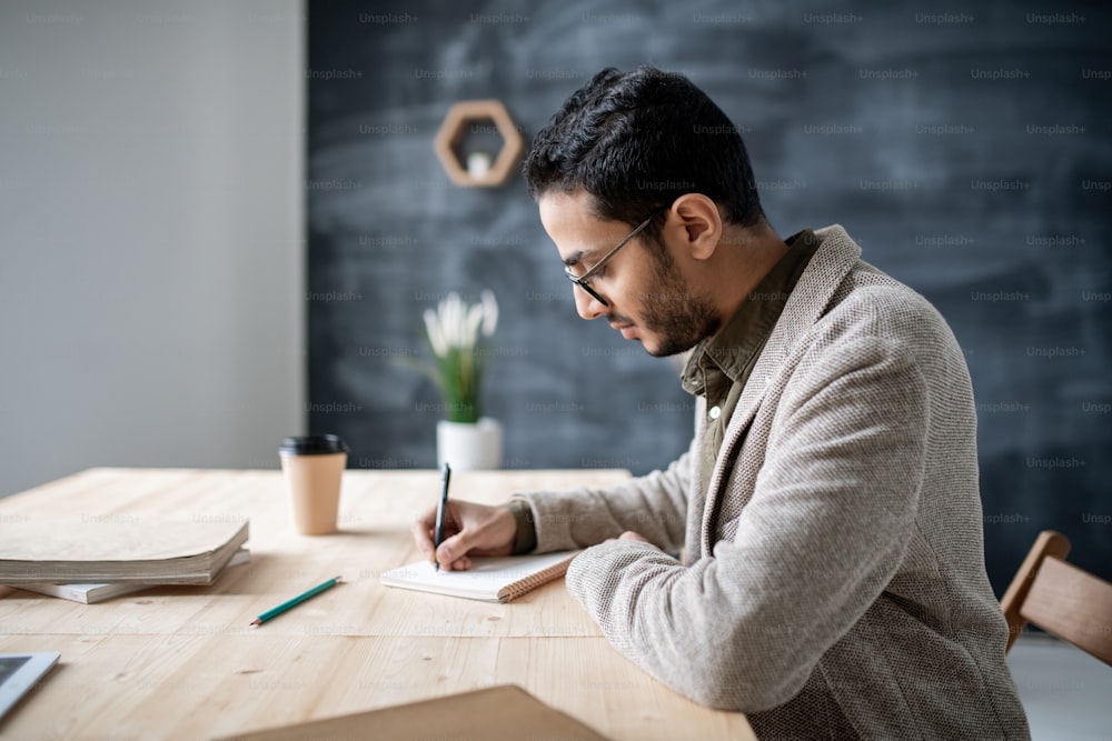 Young casual student making notes in notebook while sitting by wooden table and getting ready with homework