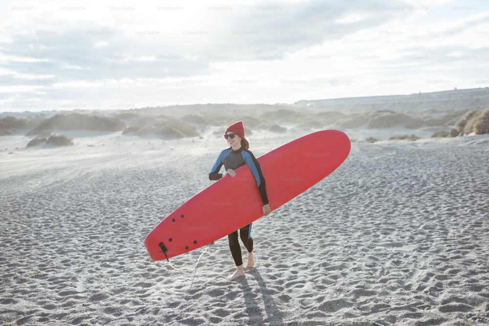 風の強いビーチで赤いサーフボードを持って歩く水着を着た若い女性サーファー。アクティブなライフスタイルとサーフィンのコンセプト