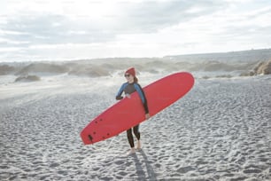 Jovem surfista mulher em maiô andando com prancha de surf vermelha na praia ventosa. Estilo de vida ativo e conceito de surf