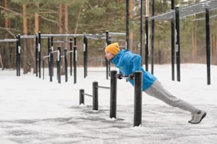 Hombre joven en chaqueta usando barra horizontal baja para practicar dominadas en el campo deportivo en invierno