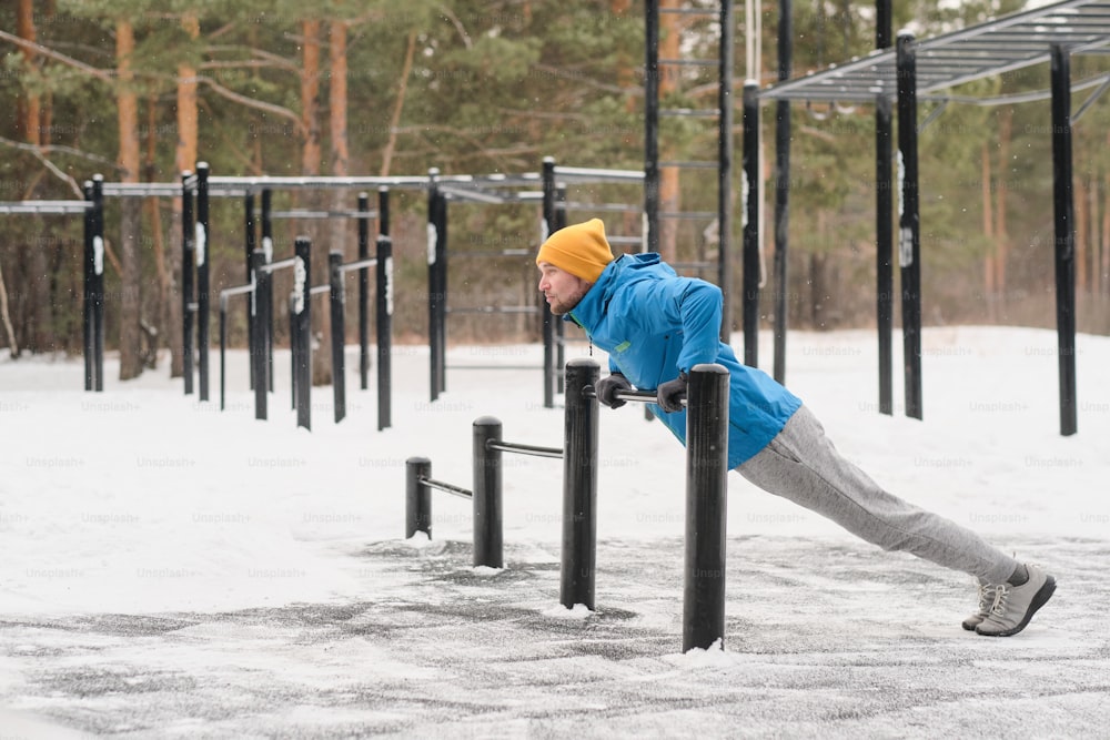 Young man in jacket using low horizontal bar to practice pull-ups on sports ground in winter
