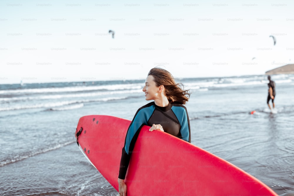 Ritratto di una giovane donna surfista in costume da bagno in piedi con tavola da surf rossa sulla spiaggia. Stile di vita attivo e concetto di surf