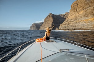 Donna che si gode il viaggio nell'oceano seduta con il salvagente sul naso dello yacht mentre naviga vicino alla costa rocciosa mozzafiato su un tramonto. Concetto di un'attività ricreativa estiva di lusso e di viaggio