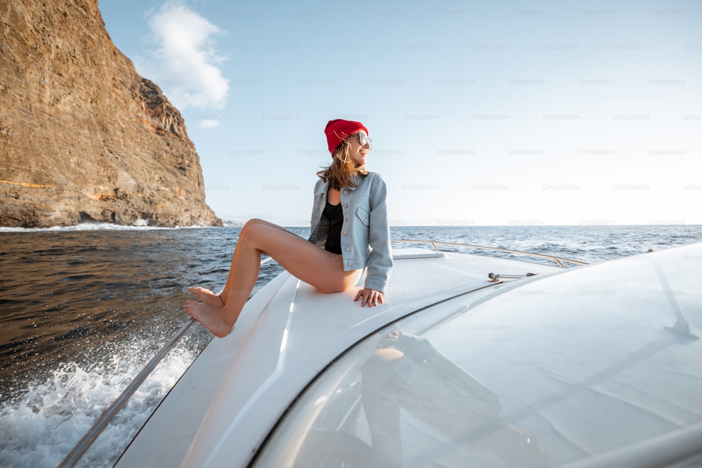 Woman dressed casually enjoying ocean voyage, sailing on a yacht near the breathtaking rocky coast on a sunset. Concept of a carefree lifestyle and travel