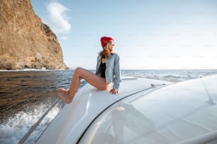 Woman dressed casually enjoying ocean voyage, sailing on a yacht near the breathtaking rocky coast on a sunset. Concept of a carefree lifestyle and travel