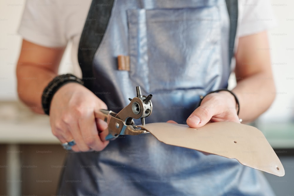 Primer plano de un trabajador irreconocible en delantal perforando agujeros en una pieza de cuero con una herramienta rotativa