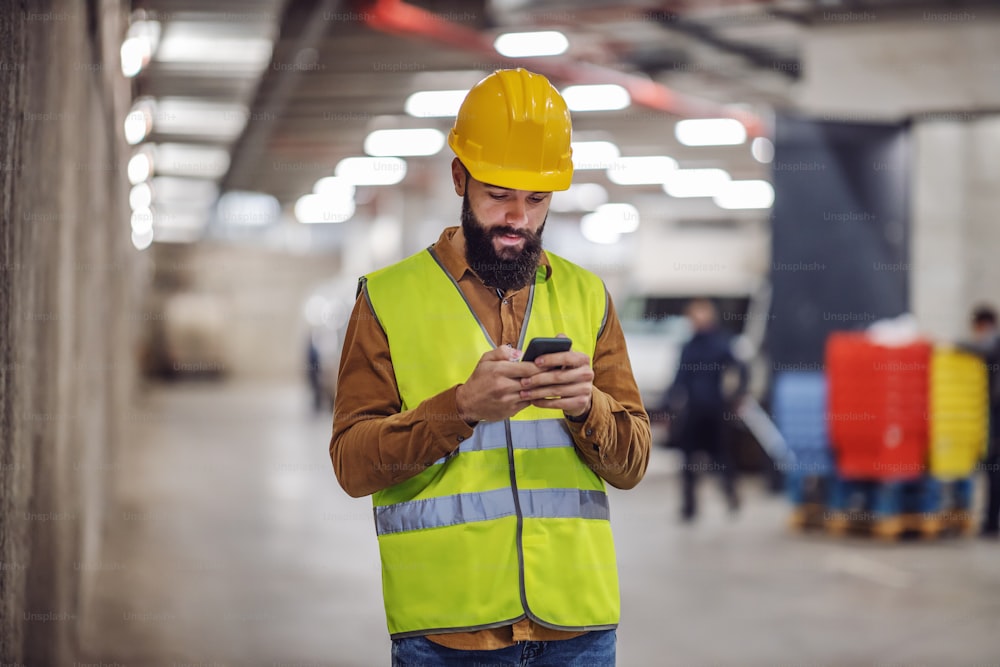 Young lazy bearded worker in vest, with helmet on head standing inside of building in construction process and hanging on social media.
