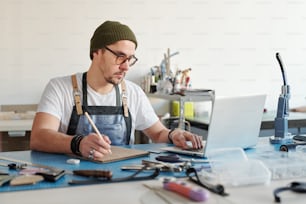 Jeune maroquinier occupé en chapeau de hipster à l’aide d’un ordinateur portable tout en planifiant l’approvisionnement en matériaux en cuir dans l’atelier