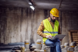 Jovem atraente supervisor barbudo focado em colete com capacete na cabeça em pé dentro do estacionamento subterrâneo em processo de construção e usando laptop.