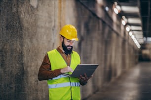 Handsome bearded supervisor in vest with helmet on head standing in building in construction process and using laptop to check is everything according to plan.