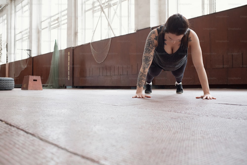 Core strengthening exercise. Low angle view of young Caucasian woman with tattooed arm doing plank in sports hall