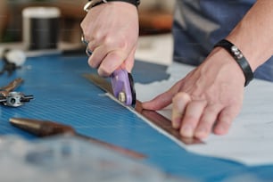 Close-up of unrecognizable man using cutting wheel and metal ruler while producing leather goods at table