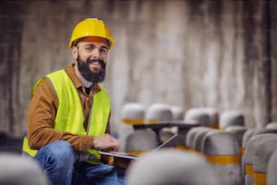 Good-looking smiling bearded supervisor in vest with hardhat on head crouching, holding laptop and looking at camera. Tunnel in construction process interior.