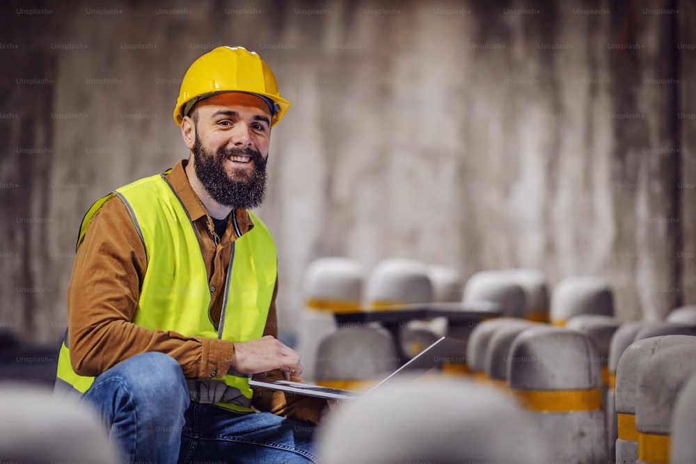 Good-looking smiling bearded supervisor in vest with hardhat on head crouching, holding laptop and looking at camera. Tunnel in construction process interior.