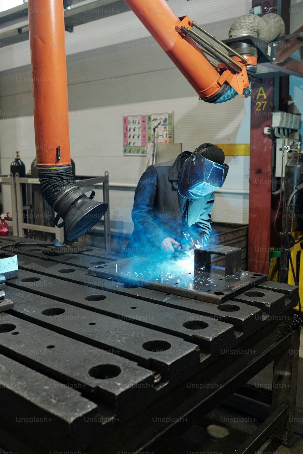 Professional welder in workwear and protective mask welding parts of huge iron industrial machine in factory workshop