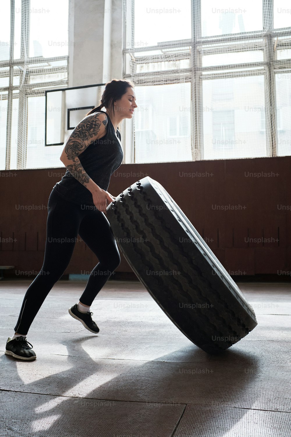 Entraînement croisé. Vue en contre-plongée d’une jeune femme sportive tatouée en vêtements de sport noirs pour développer ses muscles en retournant un pneu dans un gymnase