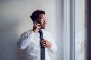 Portrait of attractive african american businessman standing next to window and looking trough it while having a phone conversation with colleague.