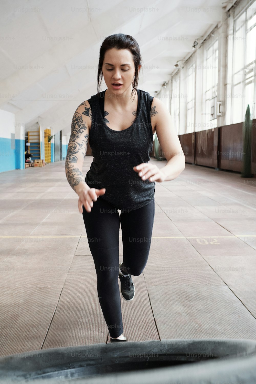 Entraînement de retournement de pneu. Jeune femme tatouée caucasienne en vêtements de sport noirs obtenant un pneu pour faire des exercices d’entraînement croisé dans le gymnase