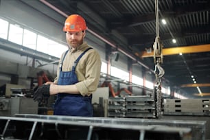 Bearded engineer in workwear and hardhat standing by huge iron detail while controling working process in factory