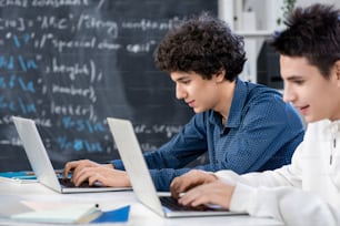 Two university students sitting by desk in front of laptops while scrolling through online information for their project