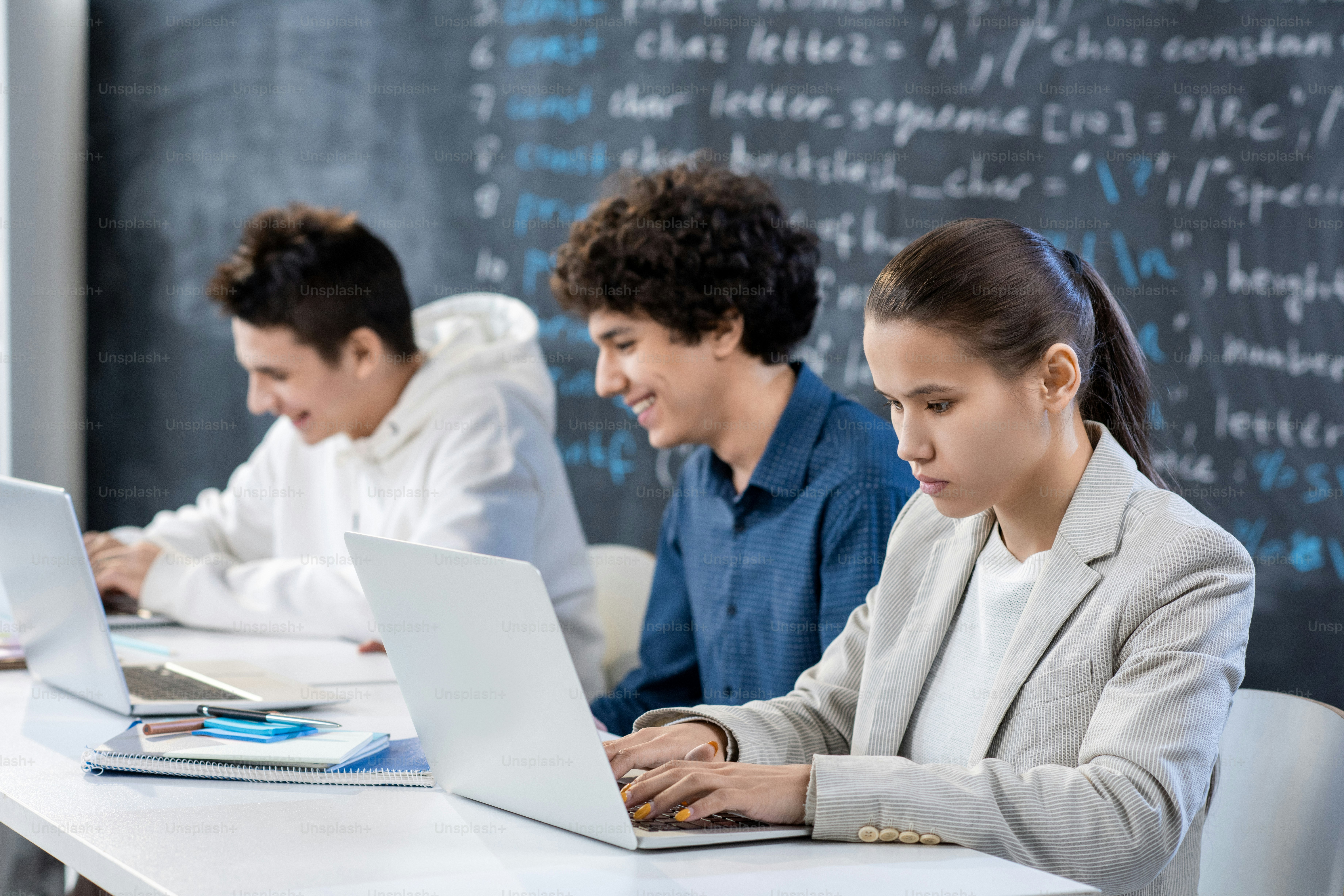 Row of young contemporary university students co-working by desk in front of laptops on background of blackboard