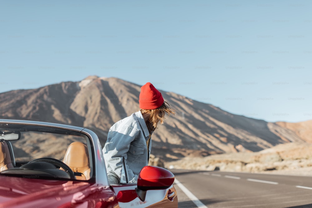 사막 계곡에서 카브리올레로 여행하는 빨간 모자를 쓴 여자, 배경에 그림 같은 화산 근처 길가에서 차에서 내린다