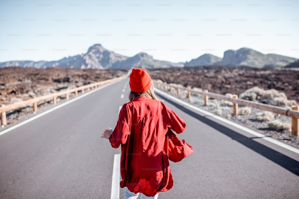 Woman stylishly dressed in red walking on the beautiful road in the midst of volcanic valley, view from the backside. Carefree lifestyle and travel concept