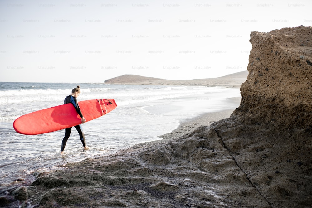 夕暮れ時に美しい岩だらけの海のビーチを赤いサーフボードで歩く女性。ウォータースポーツとアクティブなライフスタイルのコンセプト