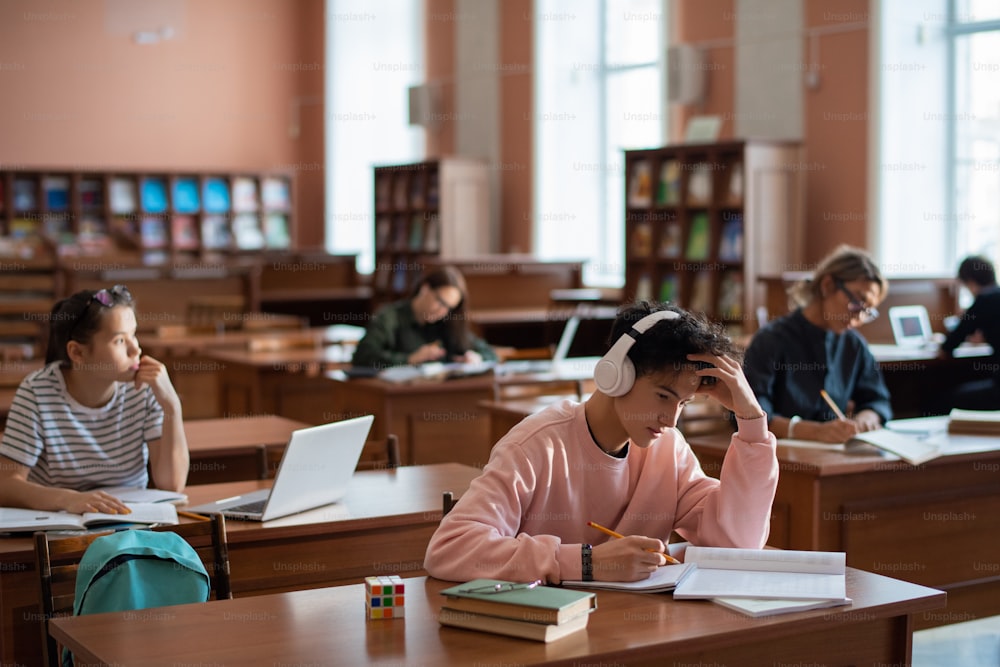 Gruppe zeitgenössischer College-Lernender, die an Schreibtischen in der Bibliothek sitzen und sich individuell auf Seminar oder Unterricht vorbereiten