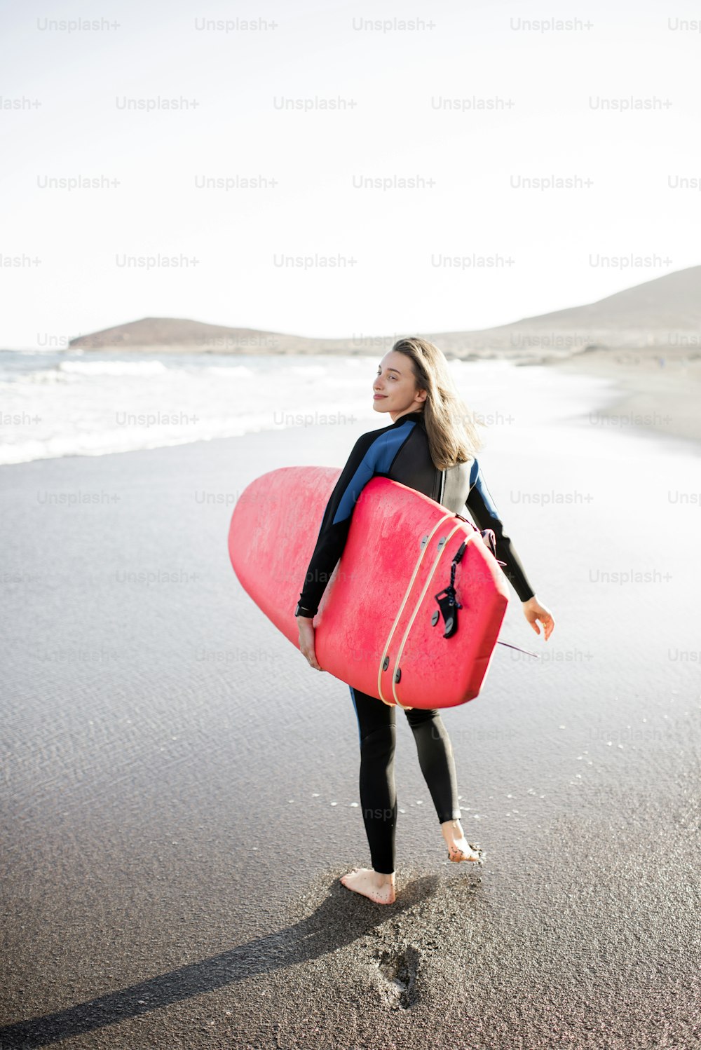 Mujer joven en traje de neopreno caminando con tabla de surf, dejando huellas en la arena detrás, vista desde la parte trasera. Concepto de deporte acuático y estilo de vida activo