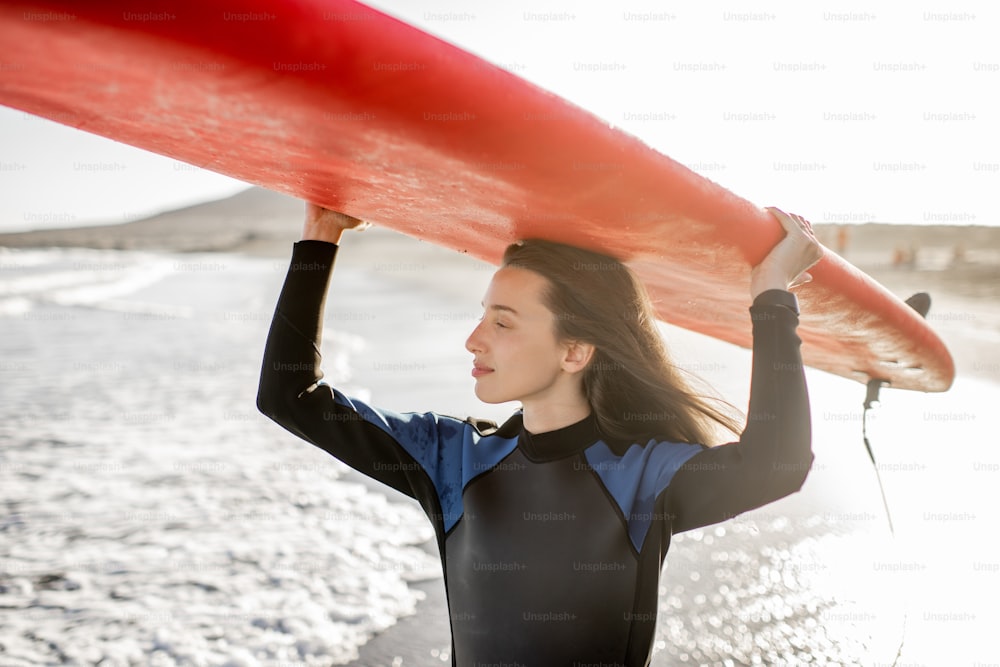 Retrato de una mujer joven en traje de neopreno que lleva el surboard por encima de la cabeza, preparándose para surfear en la playa salvaje en una puesta de sol. Concepto de estilo de vida activo