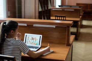 Ragazza adolescente pensierosa seduta vicino alla scrivania davanti al display del computer portatile con la homepage del sito Web di apprendimento a distanza