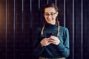 Smiling teenage girl standing indoors and typing message on social media over smart phone.