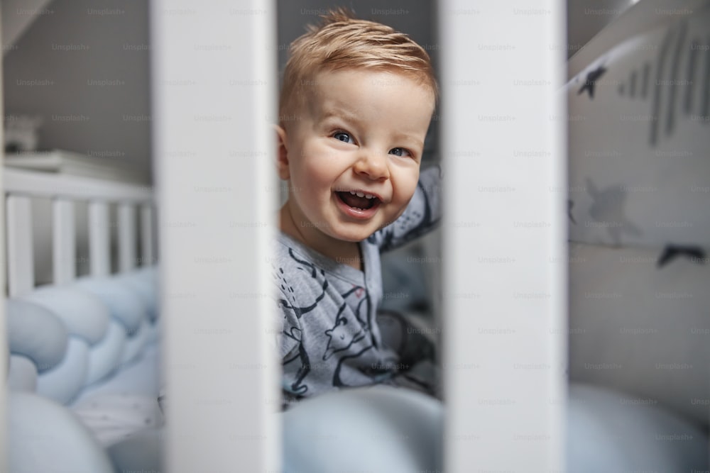 Adorable laughing blonde little baby boy with beautiful blue eyes sitting in his crib in the morning and looking at his parent.