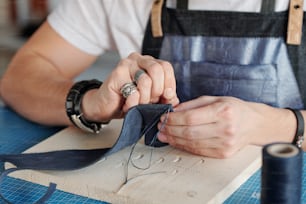 Maestro artigianale creativo con ago che tiene un piccolo pezzo di pelle scamosciata nera su una tavola di legno sul tavolo mentre cuce qualcosa