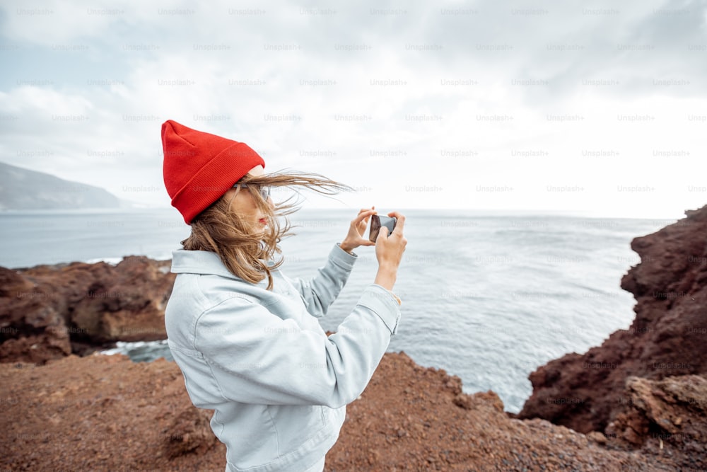 Jovem de chapéu vermelho desfrutando de uma viagem em uma costa rochosa do oceano, fotografando ao telefone paisagens de tirar o fôlego. Viajando na ilha de Tenerife, Espanha