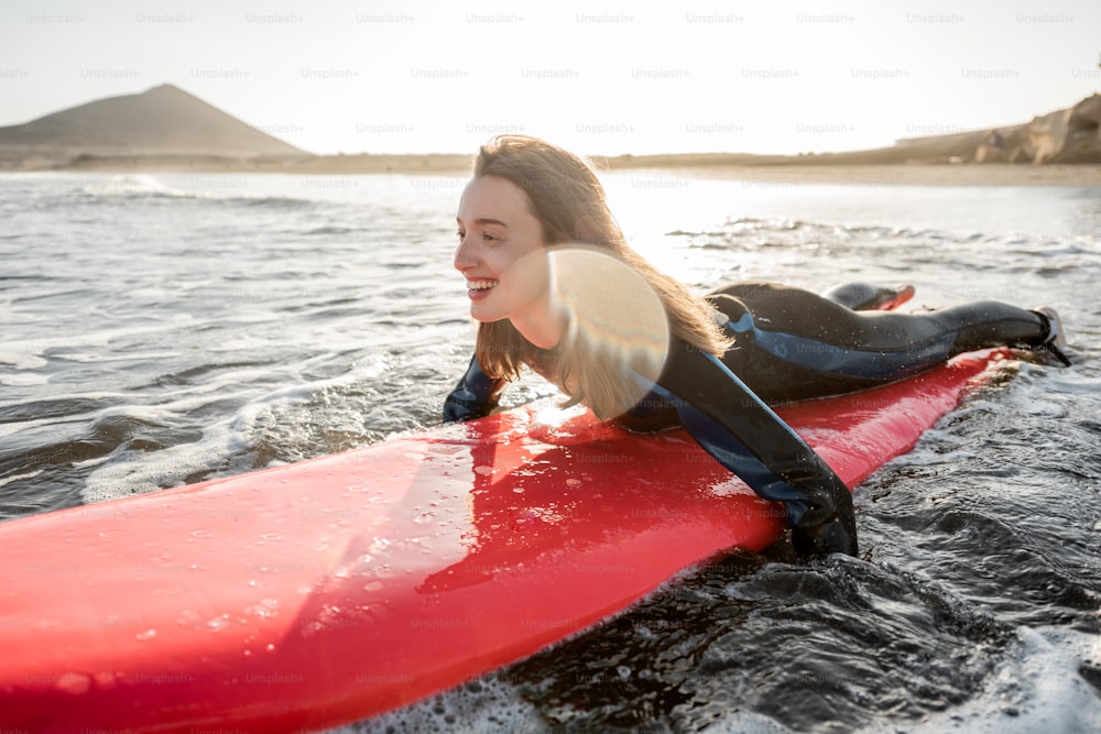 Mujer joven en traje de neopreno que atrapa el flujo de agua en la tabla de surf, surfeando en el océano ondulado durante una puesta de sol. Concepto de deportes acuáticos y estilo de vida activo