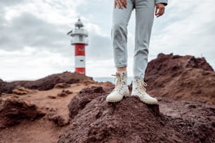 Traveler standing on the rocky land, traveling volcanic landsacpes near the lighthouse. View on the woman's trekking shoes