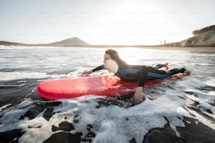 Young woman in wetsuit catching water flow on the surfboard, surfing on the wavy ocean during a sunset. Water sports and active lifestyle concept