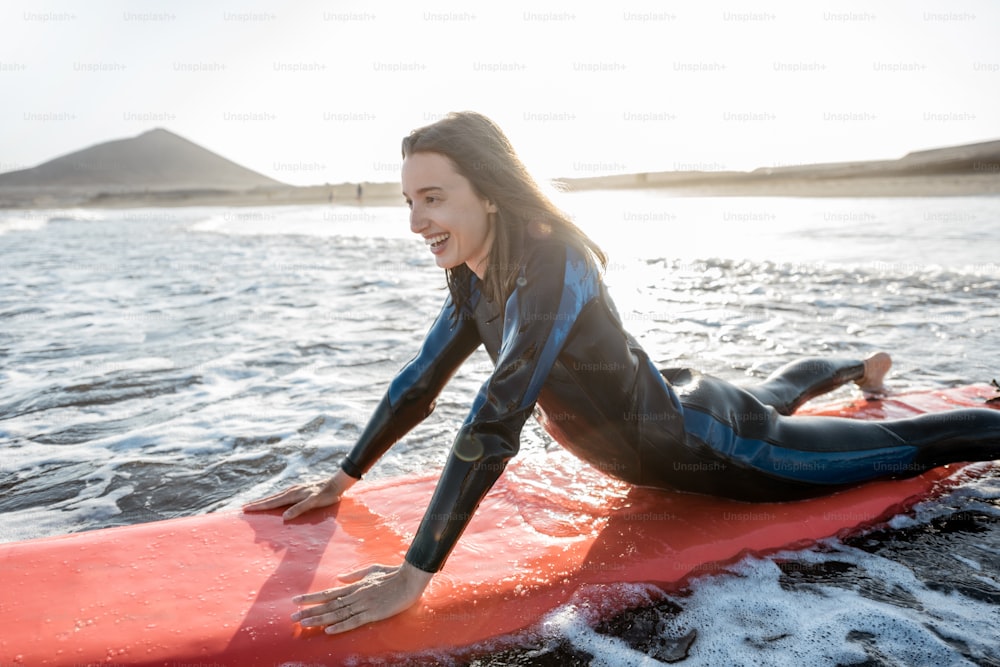 잠수복을 입은 젊은 여자는 서핑 보드에서 물의 흐름을 잡고, 일몰 동안 물결 모양의 바다에서 서핑을 합니다. 수상 스포츠와 활동적인 라이프 스타일 개념