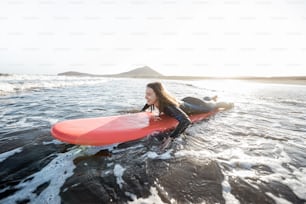 Mulher jovem em roupa de mergulho pegando o fluxo de água na prancha de surf, surfando no oceano ondulado durante um pôr do sol. Conceito de desportos aquáticos e estilo de vida ativo