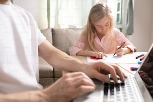 Cute blond little girl sitting by table and drawing picture on paper with highlighter while her father working in the net on quarantine