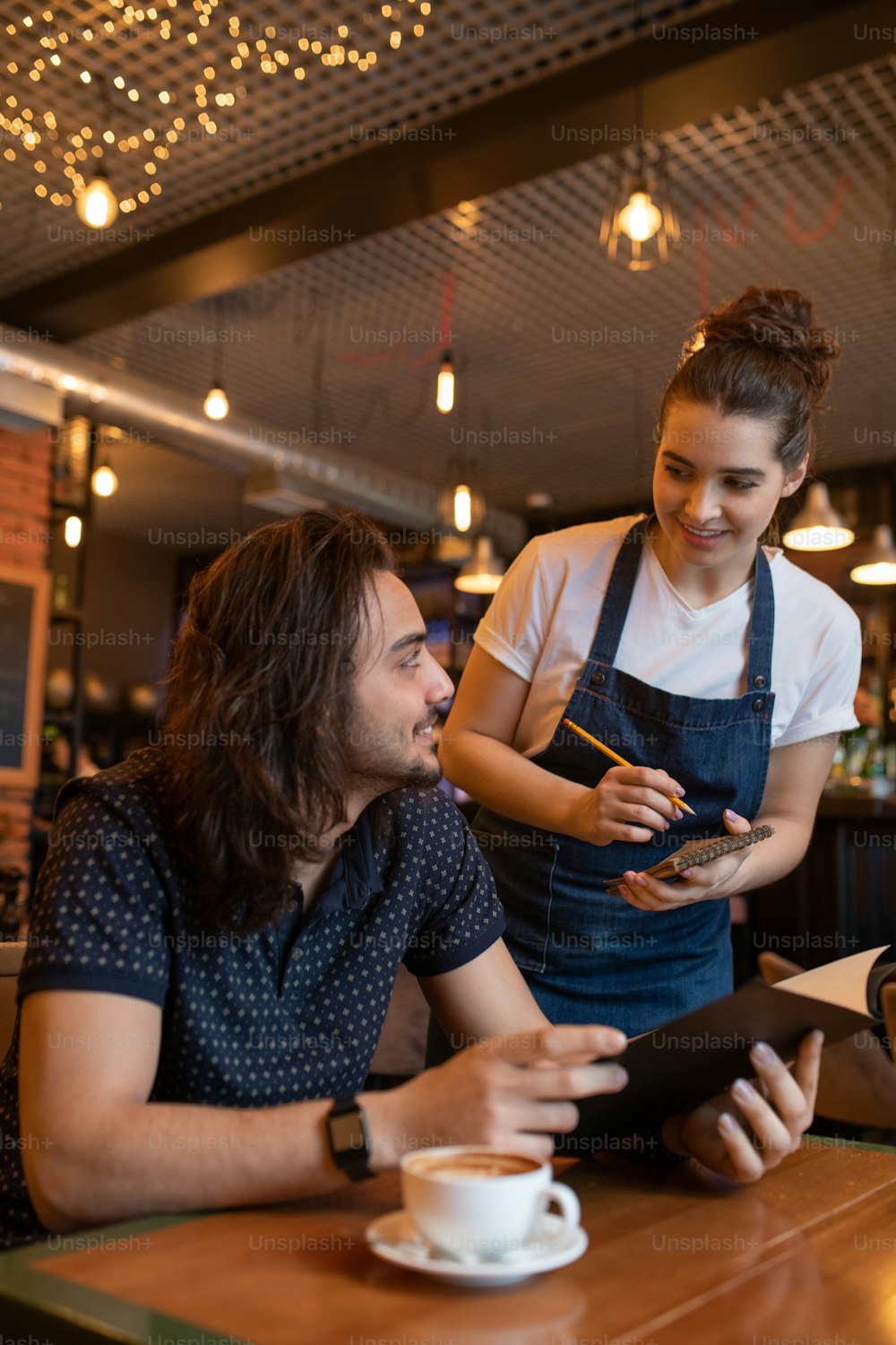 Happy young waitress in apron bending over one of clients of cafe or restaurant while writing down his order in notepad