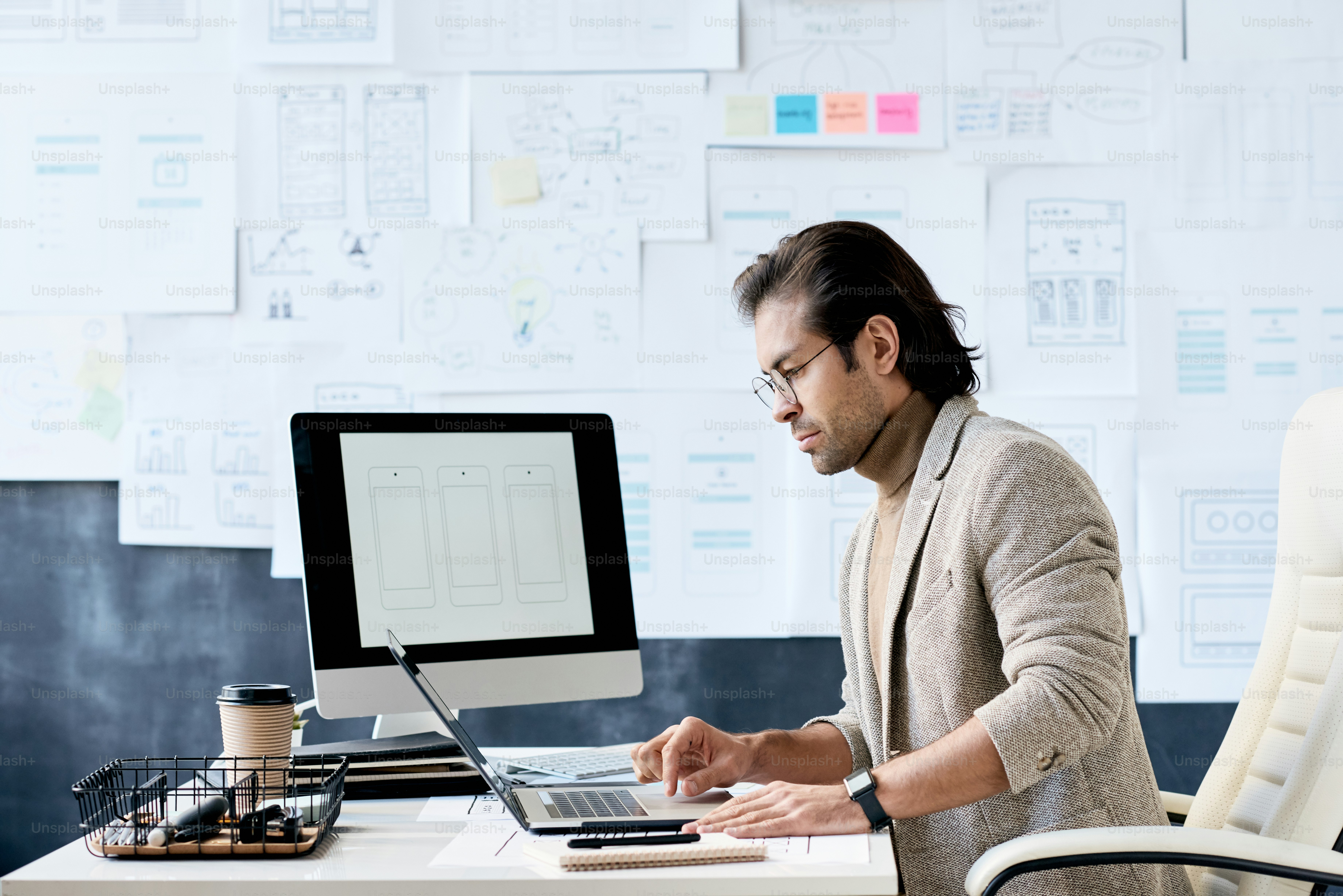 Seriöser Manager der App-Design-Firma, der mit Plantafel am Tisch im Büro sitzt und den Laptop für die Arbeit benutzt