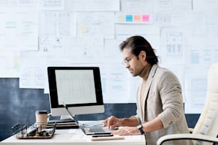 Serious manager of app design company sitting at table in office with planning board and using laptop for work