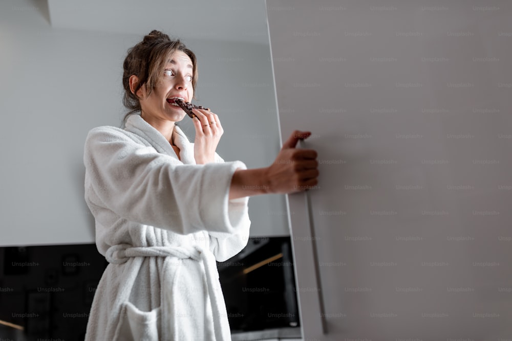 Young woman in bathrobe eating chocolate near the fridge, feeling hungry at night. Concept of not regular eating and overeating at night