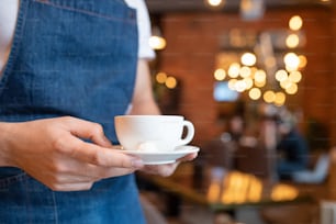 Hands of young waiter in blue apron carrying cup of coffee or tea on white porcelain saucer for one of clients of cafe or restaurant